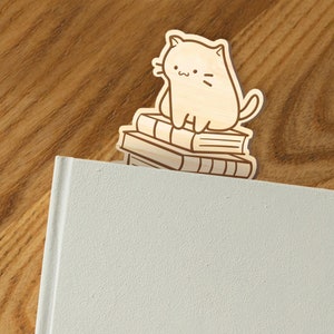 May include: Wooden bookmark featuring a cartoon cat sitting on a stack of books. The bookmark is light brown and the cat is white with black eyes and a black nose.