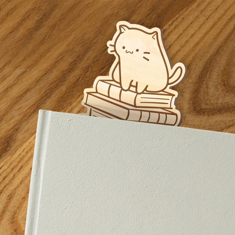 May include: Wooden bookmark featuring a cartoon cat sitting on a stack of books. The bookmark is light brown and the cat is white with black eyes and a black nose.