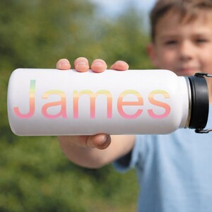 May include: White insulated water bottle with the name "James" in a gradient of pastel colors. The bottle has a black cap and a silver handle. The background is a blurred outdoor scene.