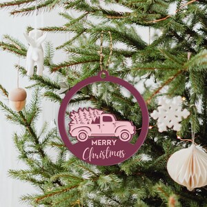 May include: A pink wooden Christmas ornament shaped like a circle with a pink truck carrying a Christmas tree inside. The ornament has the words "Merry Christmas" written on it.