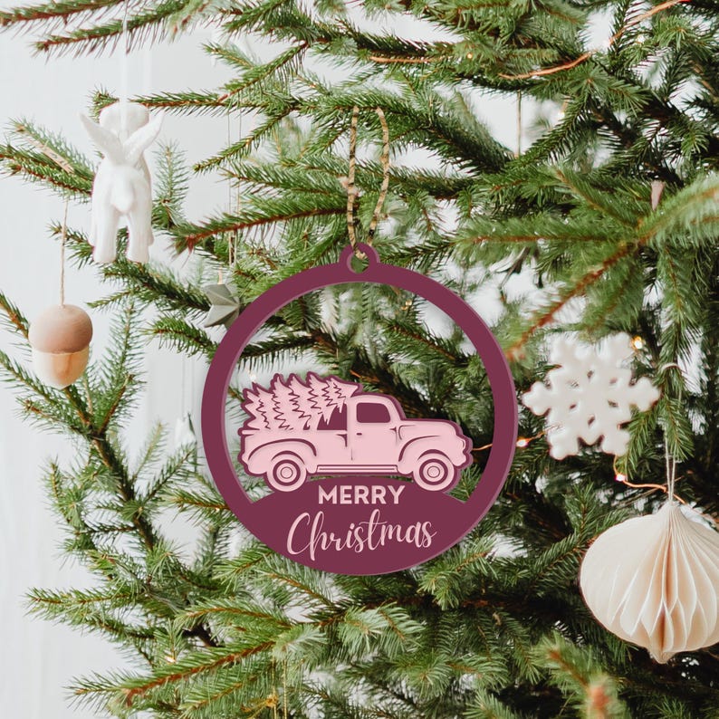 May include: A pink wooden Christmas ornament shaped like a circle with a pink truck carrying a Christmas tree inside. The ornament has the words "Merry Christmas" written on it.