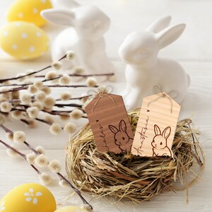 May include: Two wooden Easter bunny gift tags with twine bows. The tags are brown and natural wood and feature a bunny silhouette with the names "Chloe" and "Anastasia" engraved on them. The tags are nestled in a bird's nest.