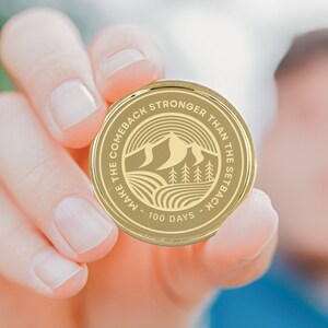 May include: Gold-colored coin with the text "Make the comeback stronger than the setback - 100 days" and a mountain range with trees in the center.