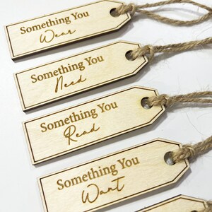 Pu&ograve; includere: Quattro etichette regalo in legno con spago attaccato. Ogni etichetta ha il testo "Something You" seguito da una parola diversa: "Wear", "Need", "Read" e "Want".
