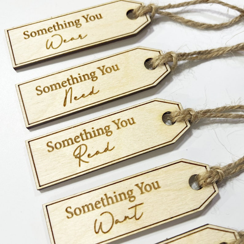 Pu&ograve; includere: Quattro etichette regalo in legno con spago attaccato. Ogni etichetta ha il testo "Something You" seguito da una parola diversa: "Wear", "Need", "Read" e "Want".