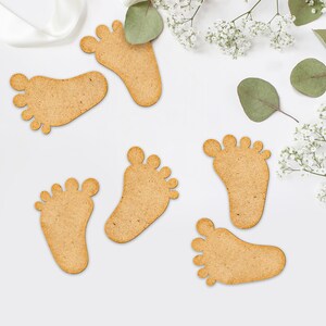 Op de afbeelding: Zes houten babyvoetjes in een lichtbruine kleur, gerangschikt op een witte achtergrond met witte bloemen en groene bladeren.