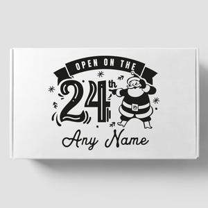 May include: Black and white illustration of Santa Claus pointing to the text "Open on the 24th" with a star and arrow design. The text "Any Name" is below the illustration.
