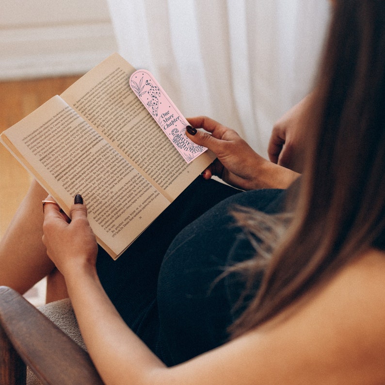 May include: A pink bookmark with a floral design and the text "One More Chapter". The bookmark is being held by a person reading a book.