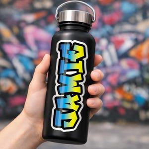 Op de afbeelding: Een zwarte waterfles met een zilveren deksel en handvat. De fles heeft een sticker met het woord "ATMWL" in een graffiti-stijl lettertype, met gele en blauwe kleurovergang en een witte omlijning.