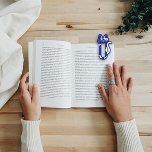 May include: A blue and white cat-shaped bookmark is placed in an open book. The book is on a wooden table with a white cloth on the left side of the table.