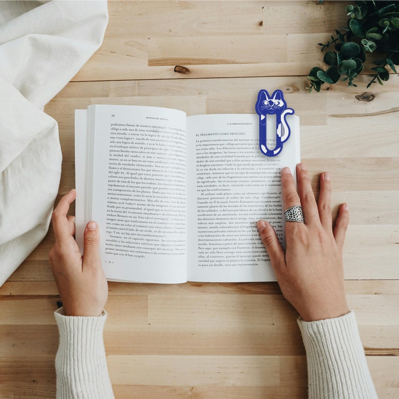 May include: A blue and white cat-shaped bookmark is placed in an open book. The book is on a wooden table with a white cloth on the left side of the table.