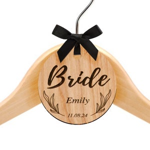 Puede incluir: Percha de madera con una etiqueta redonda grabada que dice "Bride" con el nombre "Emily" y la fecha "11.08.24". La etiqueta est&aacute; decorada con un dise&ntilde;o floral y un lazo de sat&eacute;n negro.