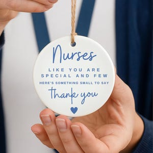 May include: A white ceramic ornament with a blue text that reads "Nurses, like you are special and few, here's something small to say thank you" with a blue heart. The ornament is held by a person.