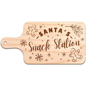 May include: Wooden cutting board with a handle, engraved with "SANTA'S Snack Station" in a decorative font. The design includes gingerbread men, snowflakes, and sprigs, perfect for holiday entertaining.