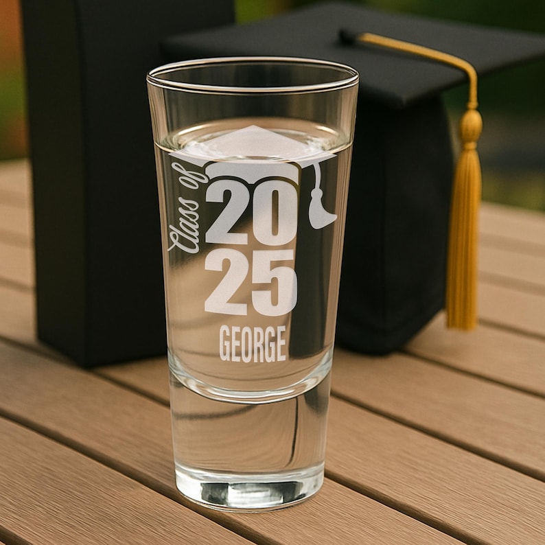 May include: A clear shot glass with the text "Class of 2025 GEORGE" in white. A graduation cap and a black box are in the background. The glass is filled with a clear liquid and sits on a wooden surface.