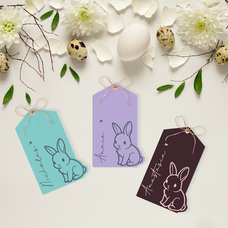May include: Three pastel colored gift tags with bunny illustrations and names written on them. The tags are tied with twine and are on a white background with white flowers and quail eggs.