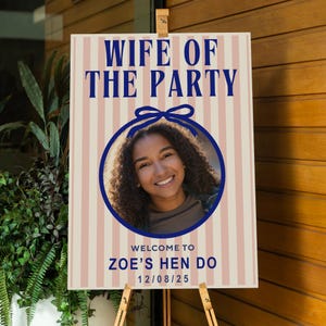 Puede incluir: Un letrero de fiesta con el texto "WIFE OF THE PARTY" en azul marino, sobre una foto de una mujer sonriente. El letrero tiene un fondo a rayas rosas y blancas y dice "WELCOME TO ZOE'S HEN DO 12/08/25". El letrero se muestra en un caballete de madera.