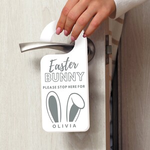 May include: White door hanger with the text "Easter Bunny" and "Please stop here for Olivia". The hanger has a gray bunny ear design.