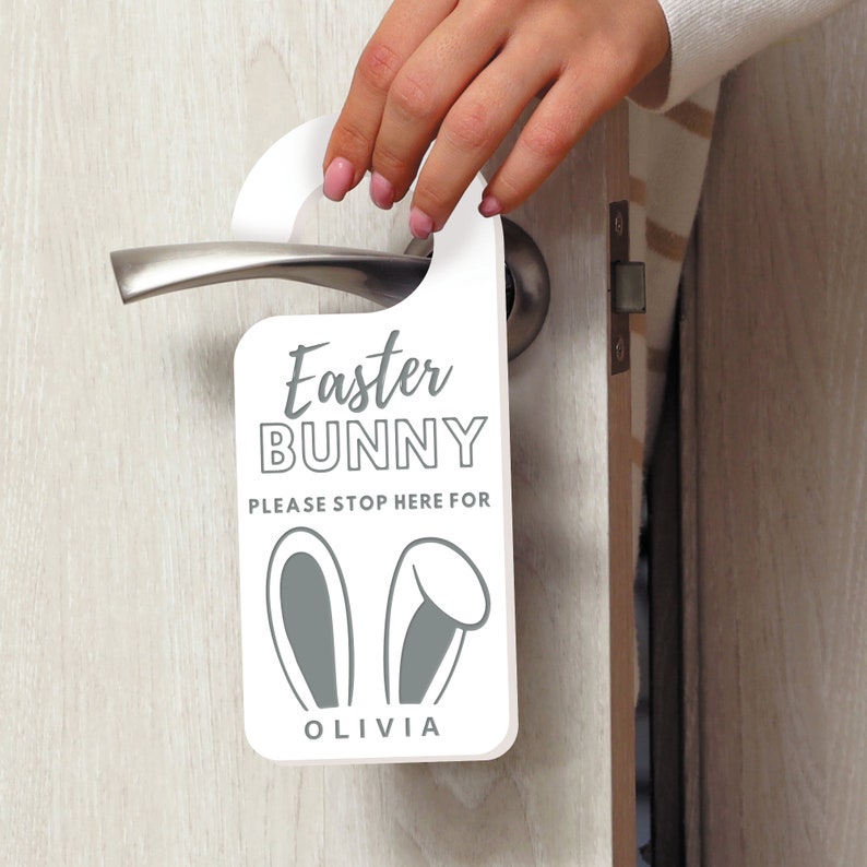 May include: White door hanger with the text "Easter Bunny" and "Please stop here for Olivia". The hanger has a gray bunny ear design.