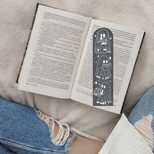 May include: A black metal bookmark featuring a ghost reading a book. The ghost is depicted in three different poses, holding a book, reading a book, and carrying a stack of books. The bookmark is a great gift for book lovers and fans of the spooky season.