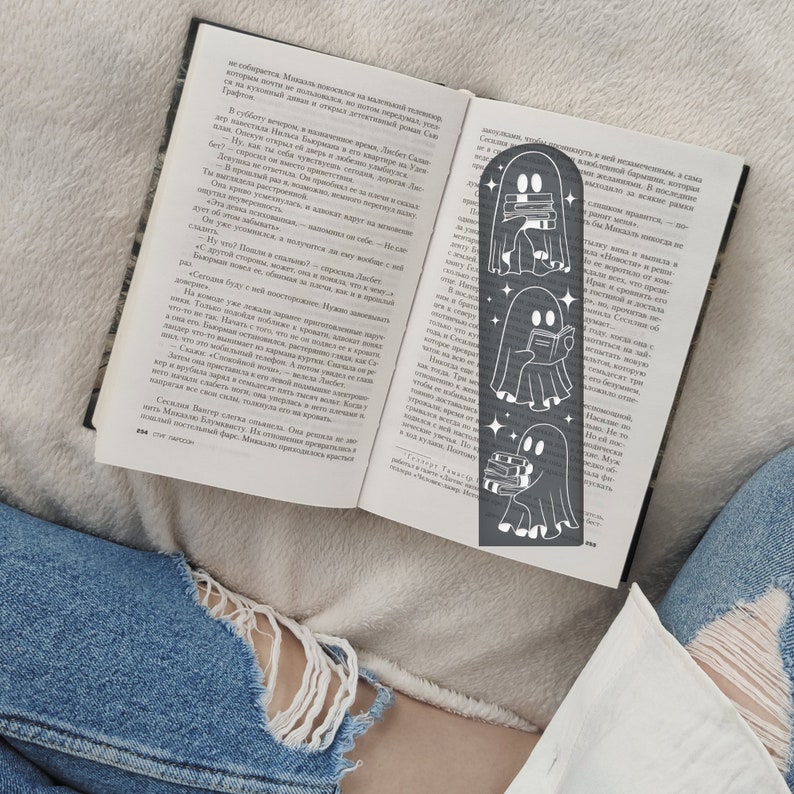 May include: A black metal bookmark featuring a ghost reading a book. The ghost is depicted in three different poses, holding a book, reading a book, and carrying a stack of books. The bookmark is a great gift for book lovers and fans of the spooky season.