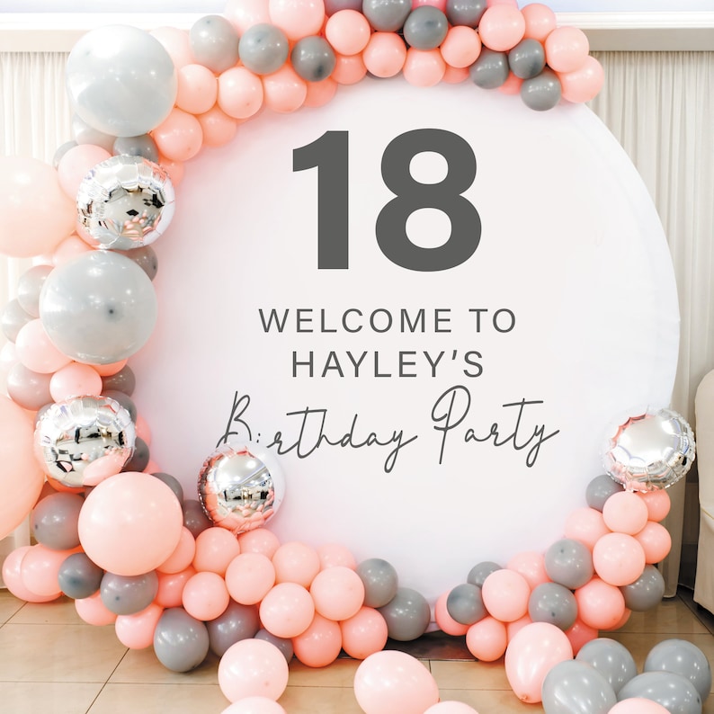 May include: A white round backdrop with the number 18 in grey text, the words "Welcome to Hayley's Birthday Party" in grey text, and a garland of pink and grey balloons.