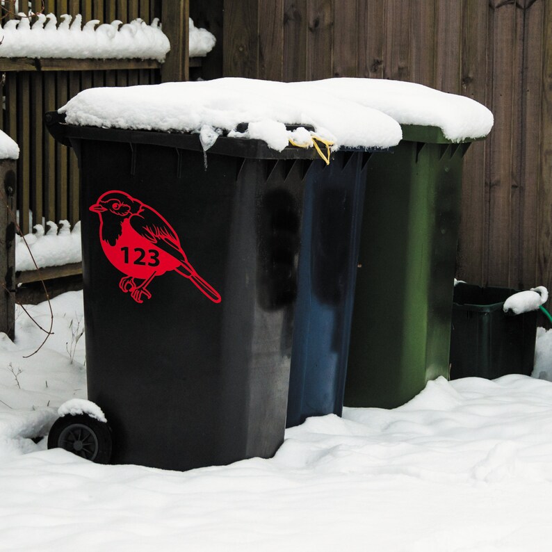May include: Three outdoor trash bins in a snowy environment. The front bin is black with a red bird graphic and the number 123. The other bins are blue and green. Snow covers the tops of the bins and the ground.