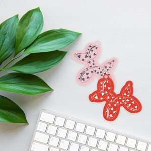 May include: Two heart-shaped balloon dog ornaments. One is pink with dark gray hearts, the other is red with white hearts. A keyboard and green leaves are in the background.