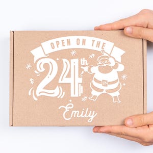 May include: A brown cardboard box with a white design featuring Santa Claus, the text "OPEN ON THE 24th", and the name "Emily".