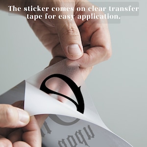 May include: A close-up of a hand peeling back a clear transfer tape from a black vinyl sticker. The sticker has the word "Happy" written in a gothic font.