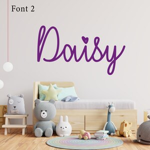 Op de afbeelding: Paarse cursieve lettertype die de naam "Daisy" spelt met een klein hartje boven de "y". De naam staat op een witte muur boven een houten bed met een grijs-wit gestreept kussen en een geel stervormig kussen.