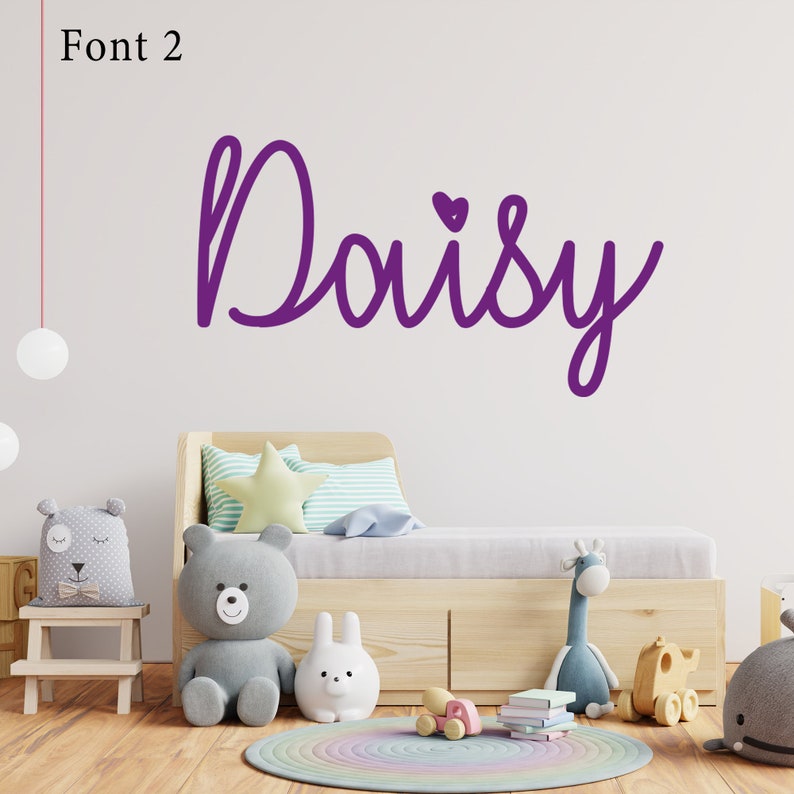 Op de afbeelding: Paarse cursieve lettertype die de naam "Daisy" spelt met een klein hartje boven de "y". De naam staat op een witte muur boven een houten bed met een grijs-wit gestreept kussen en een geel stervormig kussen.