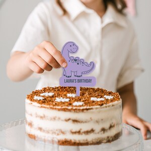 May include: Purple dinosaur cake topper with the text "Laura's Birthday" on a brown and white frosted cake.