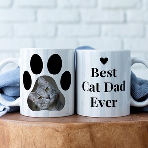May include: Two white ceramic mugs. One mug features a gray cat peeking out of a black paw print. The other mug has the text "Best Cat Dad Ever" with a black heart above the text. Both mugs have a white handle.