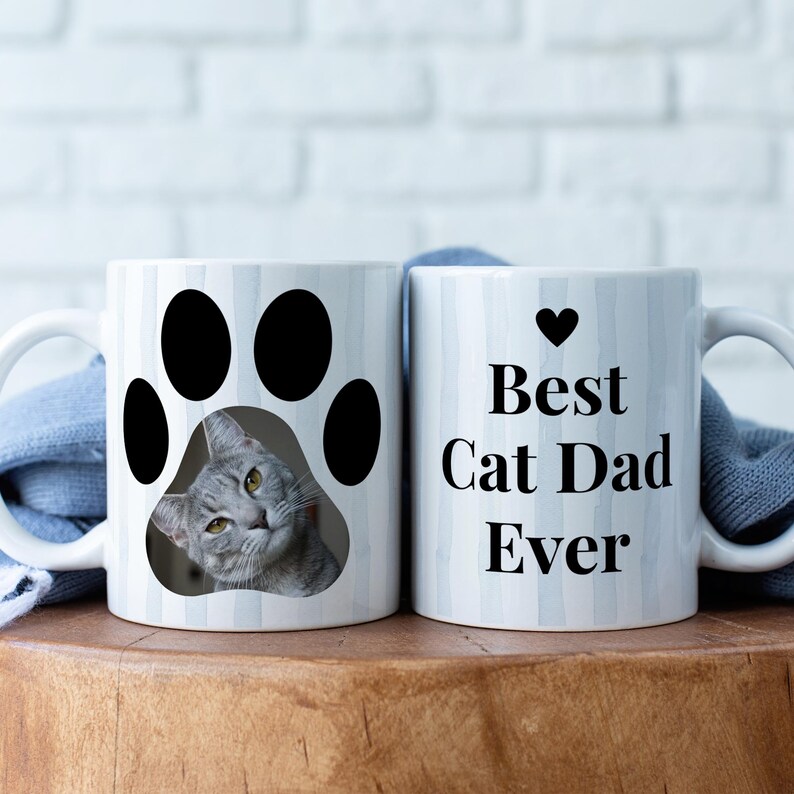 May include: Two white ceramic mugs. One mug features a gray cat peeking out of a black paw print. The other mug has the text "Best Cat Dad Ever" with a black heart above the text. Both mugs have a white handle.