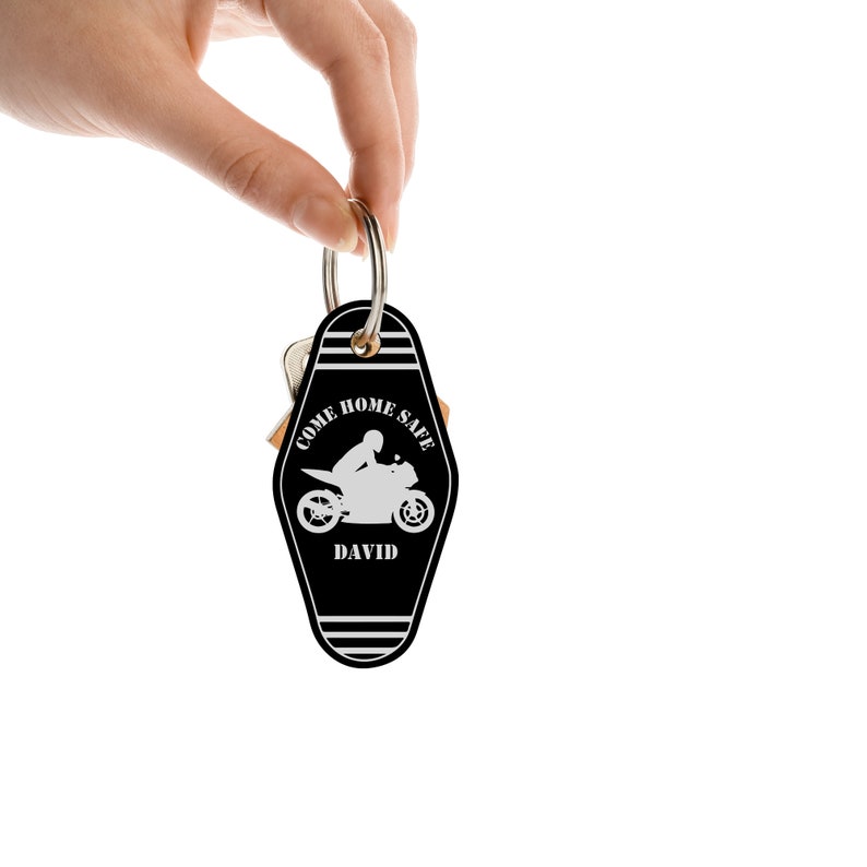 Op de afbeelding: Zwart-witte sleutelhanger met motorfiets thema, met de tekst "COME HOME SAFE" en de naam "DAVID". De sleutelhanger heeft een motorfietsafbeelding en wordt door een hand tegen een witte achtergrond gehouden.
