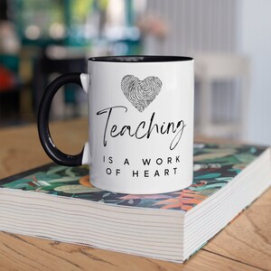 Puede incluir: Taza de cer&aacute;mica blanca con asa e interior negros. La taza presenta el texto "Teaching is a work of heart" en escritura negra, junto con un dise&ntilde;o de coraz&oacute;n de huellas dactilares. La taza est&aacute; sobre un libro con dise&ntilde;o floral.