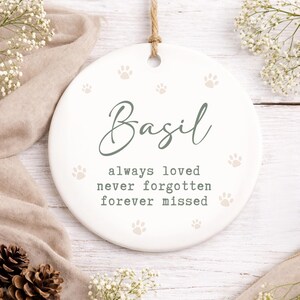 May include: A white ceramic pet memorial ornament with the name "Basil" in script, surrounded by paw prints. The text "always loved never forgotten forever missed" is below the name. A jute string is attached for hanging.
