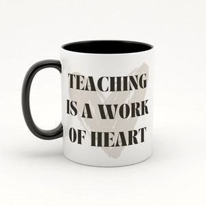 K&ouml;nnte beinhalten: Wei&szlig;e Keramiktasse mit schwarzem Rand und Henkel. Die Tasse hat ein braunes Herz mit dem Text "TEACHING IS A WORK OF HEART" darauf gedruckt.