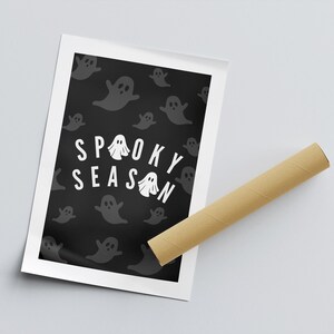 Puede incluir: Un p&oacute;ster de Halloween con la frase "Spooky Season" en blanco, rodeado de ilustraciones de fantasmas grises sobre un fondo negro. El p&oacute;ster est&aacute; enmarcado en blanco, con un tubo de cart&oacute;n marr&oacute;n al lado.