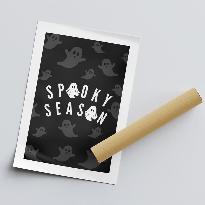 Puede incluir: Un p&oacute;ster de Halloween con la frase "Spooky Season" en blanco, rodeado de ilustraciones de fantasmas grises sobre un fondo negro. El p&oacute;ster est&aacute; enmarcado en blanco, con un tubo de cart&oacute;n marr&oacute;n al lado.