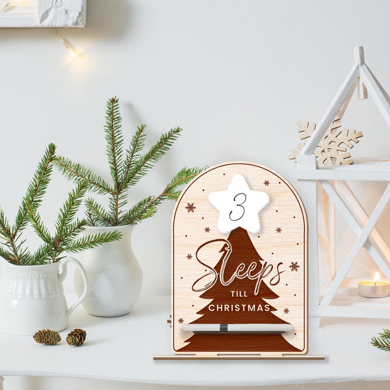 Puede incluir: Un letrero de cuenta atr&aacute;s de Navidad de madera con una estrella blanca en la parte superior que dice "3 Sleeps Till Christmas". El letrero tiene una silueta de &aacute;rbol marr&oacute;n y un marcador de borrado en seco blanco.