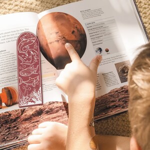 May include: A red bookmark with a white illustration of sharks and coral. The bookmark is placed on a book about Mars with a picture of the planet and facts about it. The text on the page includes "Mars Data", "Diameter: 4,212 miles (6,792 km)", "Average Distance from Sun: 141.5 million miles (227.9 million km)", "Orbital Period: 687 Earth days", "Rotation Period: 24.6 hours", "Surface Temperature: -195&deg; to 77&deg; F (-125&deg; to 25&deg; C)", "Number of Moons: 2", "Size Comparison: Mars".