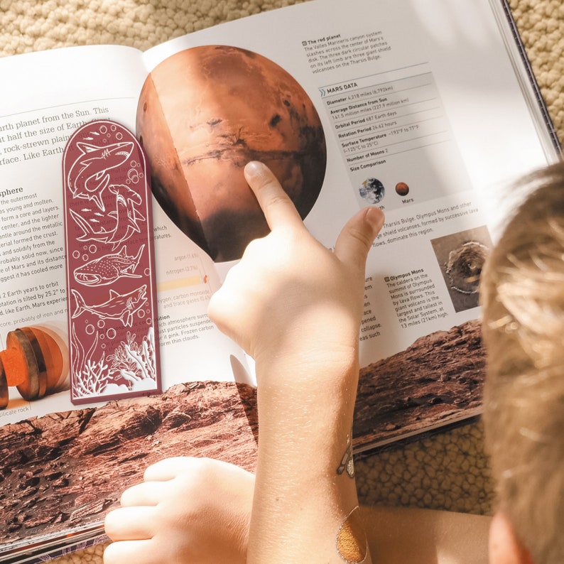 May include: A red bookmark with a white illustration of sharks and coral. The bookmark is placed on a book about Mars with a picture of the planet and facts about it. The text on the page includes "Mars Data", "Diameter: 4,212 miles (6,792 km)", "Average Distance from Sun: 141.5 million miles (227.9 million km)", "Orbital Period: 687 Earth days", "Rotation Period: 24.6 hours", "Surface Temperature: -195&deg; to 77&deg; F (-125&deg; to 25&deg; C)", "Number of Moons: 2", "Size Comparison: Mars".