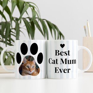 May include: Two white ceramic mugs with a cat theme. One mug features a black paw print with a photo of an orange cat. The other mug has the text "Best Cat Mum Ever" with a black heart. Both mugs have a blue and white striped pattern.