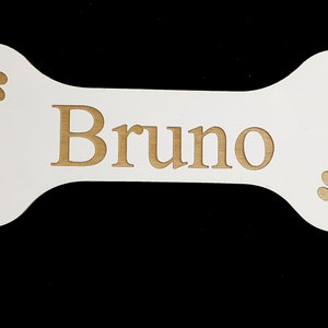 May include: White wooden bone-shaped sign with the name "Bruno" engraved in gold lettering. The sign has two gold paw prints on either side of the name.