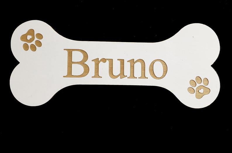 May include: White wooden bone-shaped sign with the name "Bruno" engraved in gold lettering. The sign has two gold paw prints on either side of the name.