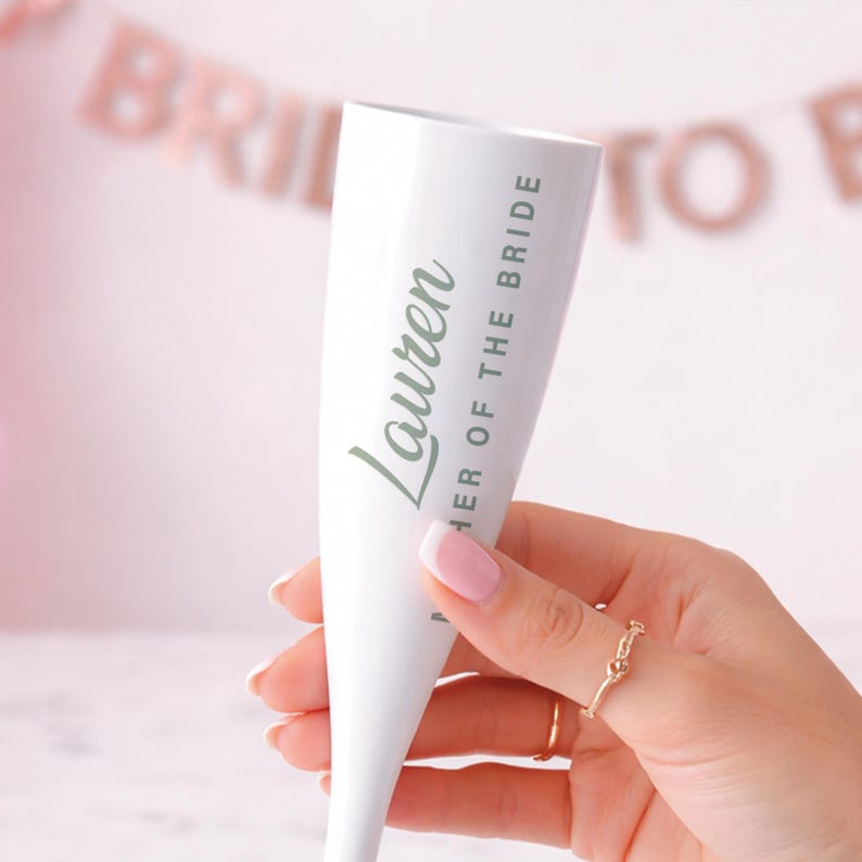 May include: A white champagne flute with the name "Lauren" and "Mother of the Bride" printed in green. The flute is held in a hand with a gold ring. A banner in the background reads "Bride to Be".