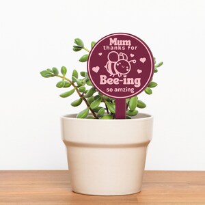 K&ouml;nnte beinhalten: Ein runder Pflanzenmarker in Burgunderrot mit einer pinkfarbenen Bienenillustration und dem Text "Mum thanks for bee-ing so amazing".