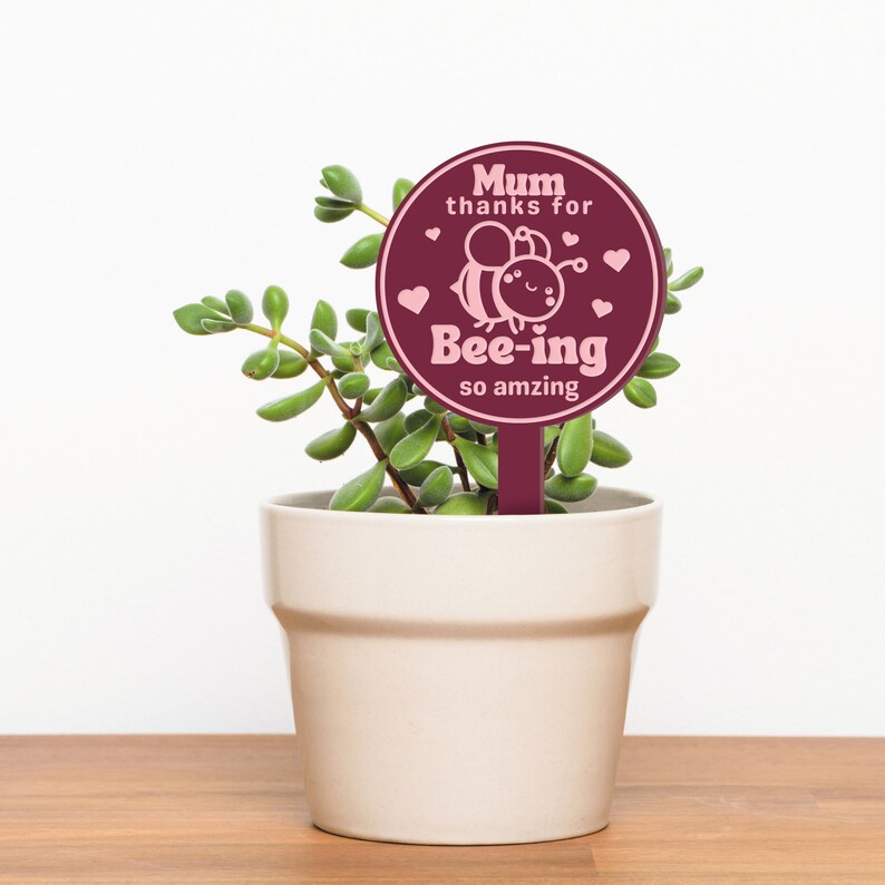K&ouml;nnte beinhalten: Ein runder Pflanzenmarker in Burgunderrot mit einer pinkfarbenen Bienenillustration und dem Text "Mum thanks for bee-ing so amazing".